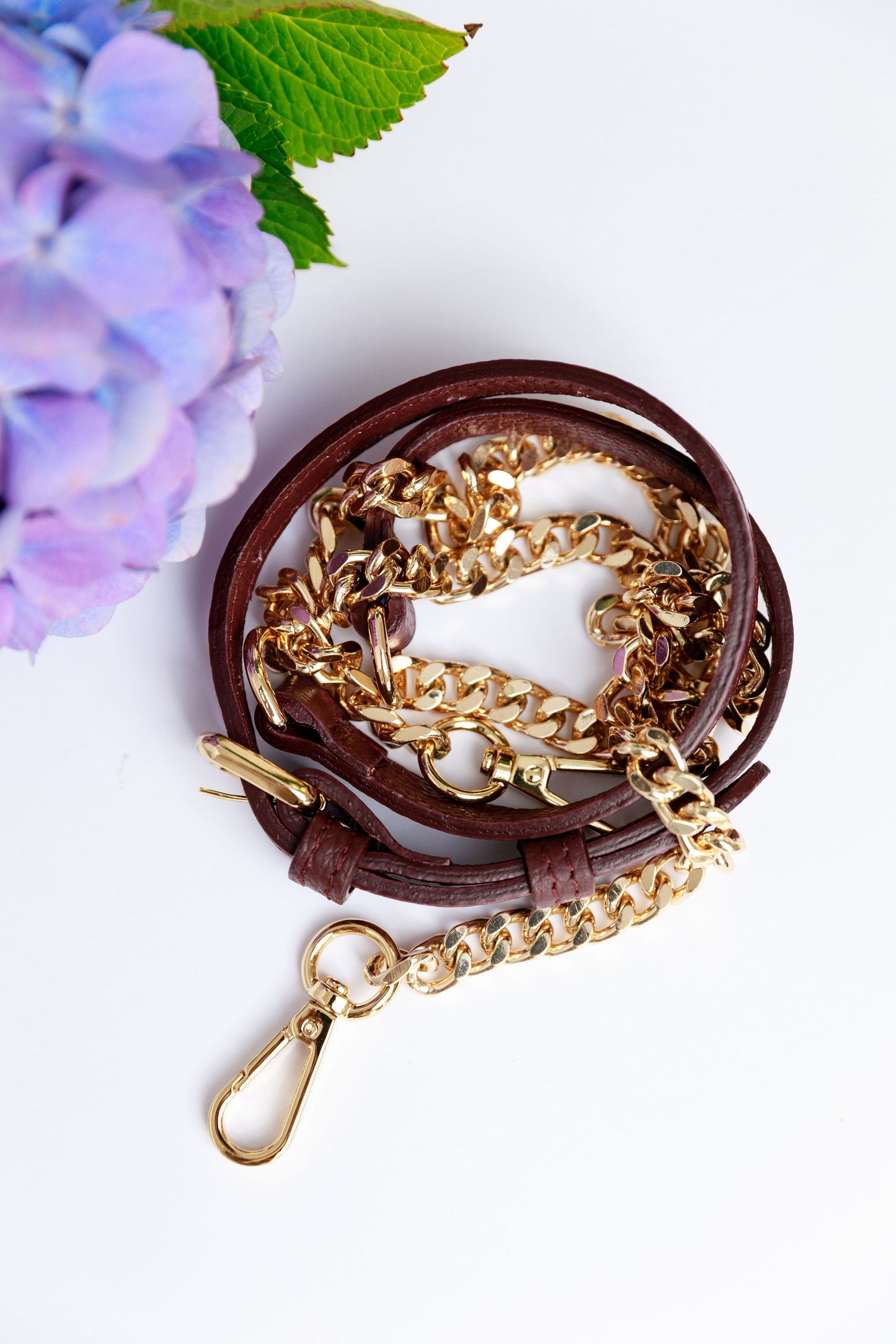 A burgundy leather and chain strap on a white surface with a blurred hydrangea in the foreground.