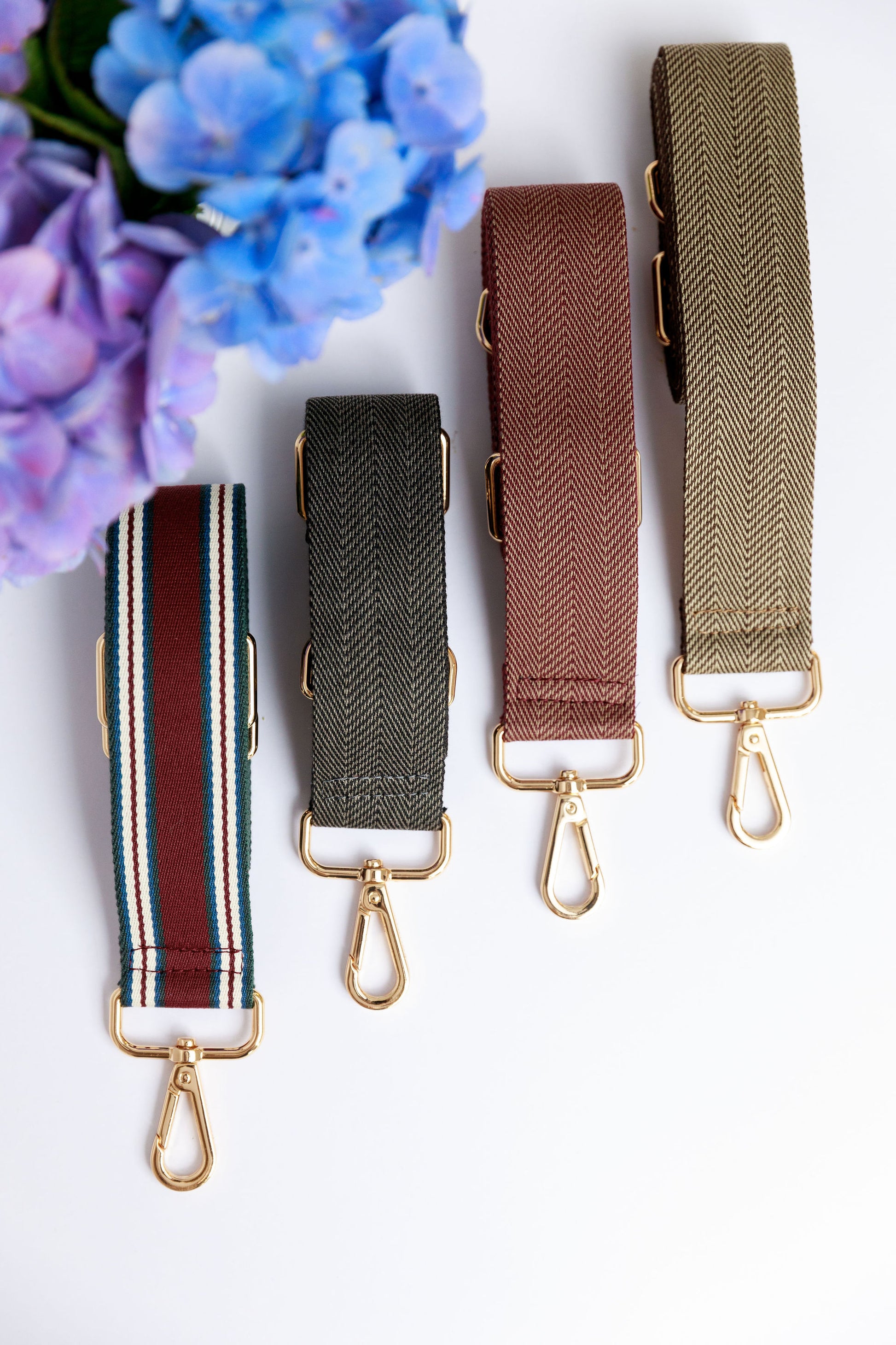 Four woven crossbody straps in burgundy, black, chocolate brown and a striped burgundy on a white surface with a blurred hydrangea in the foreground.