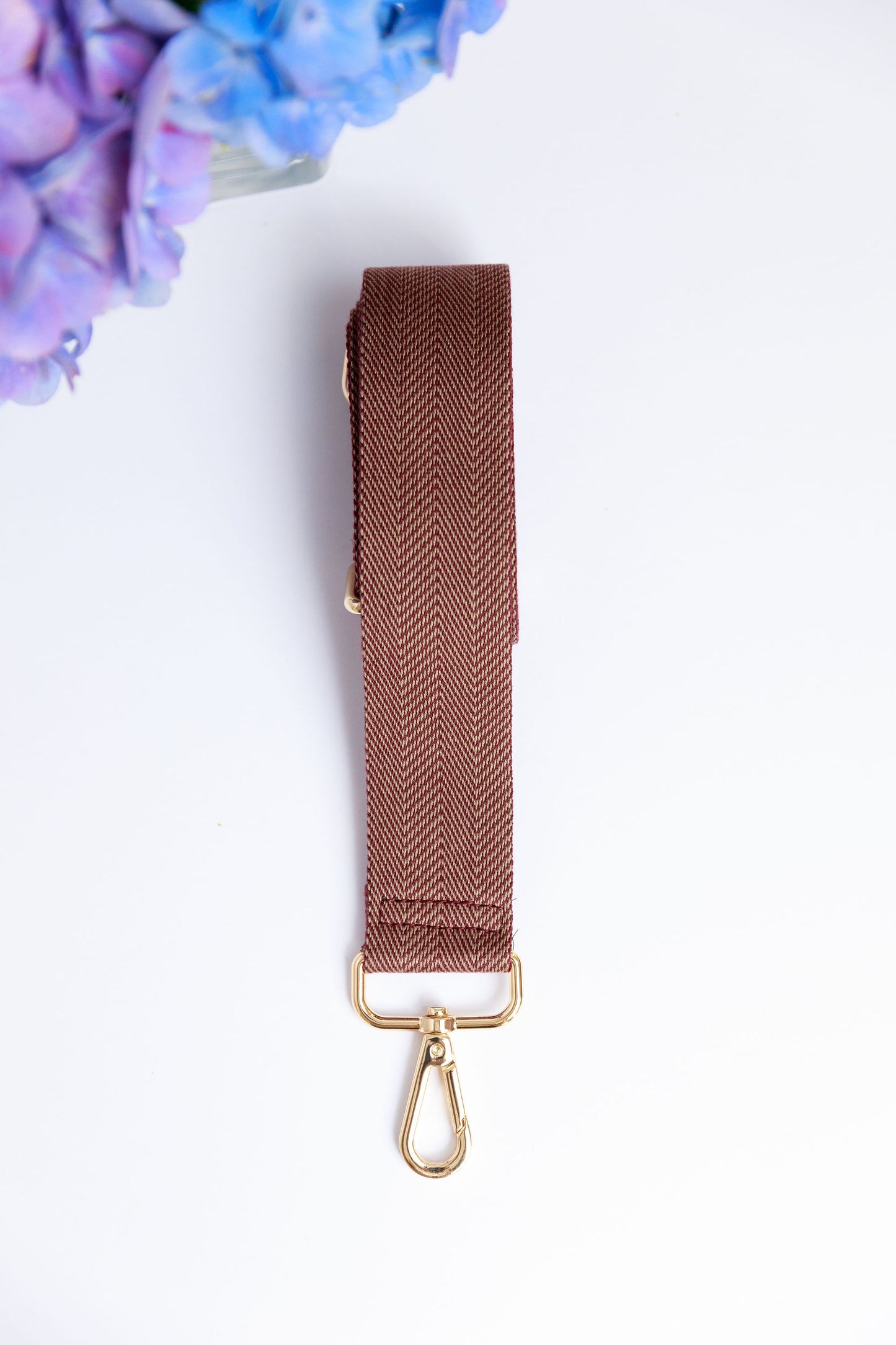 A burgundy woven strap on a white surface with a blurred hydrangea in the foreground.