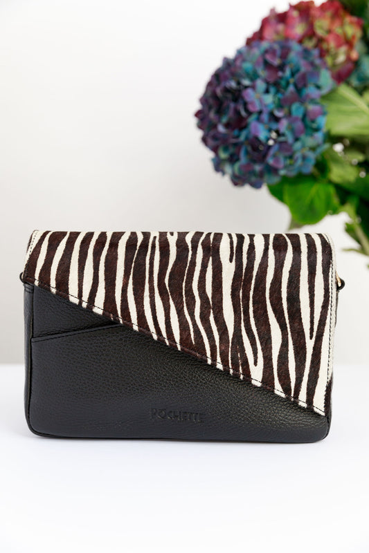 A zebra leather flap on a black leather crossbody bag on a white surface with a blurred hydrangea in the background.