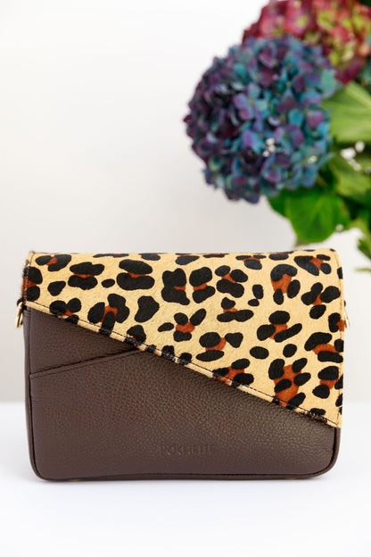 A leopard leather flap on a chocolate brown crossbody bag white surface with a blurred hydrangeas in the background.