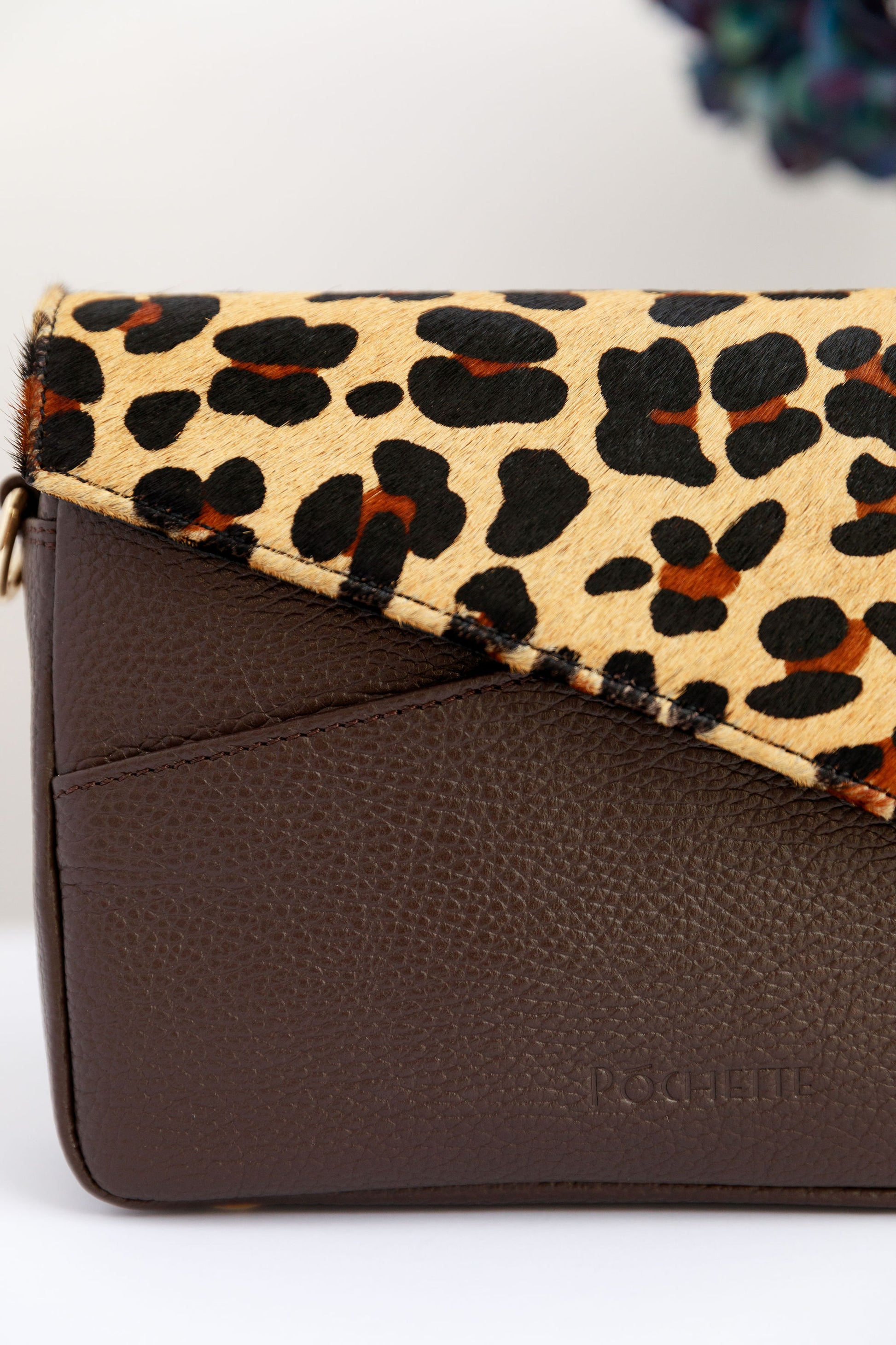 A close up of a leopard leather flap on a chocolate brown crossbody bag white surface with a blurred hydrangeas in the background.