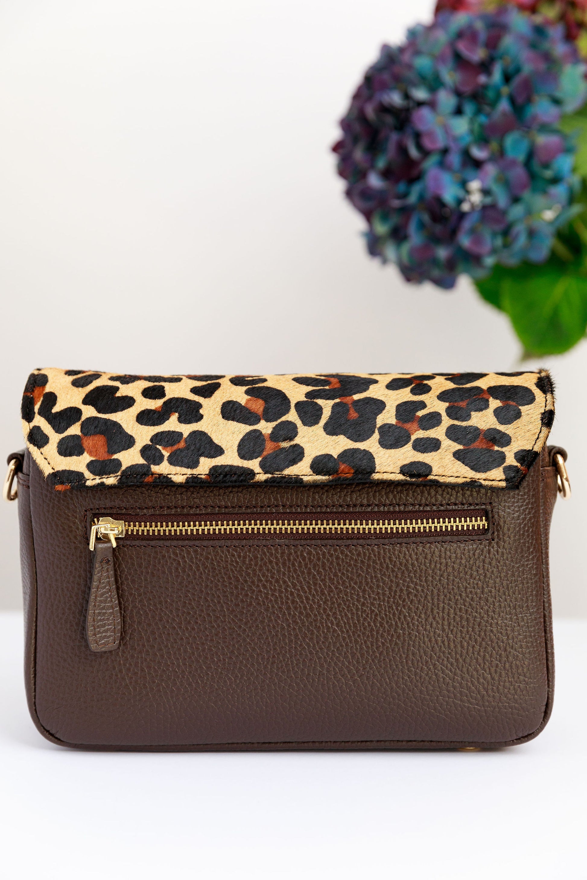 The back of a chocolate brown crossbody bag with a leopard print leather flap attached with a blurred hydrangea in the background.