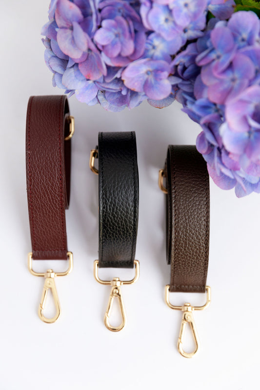 Three short leather shoulder straps in burgundy, black and chocolate brown on a white surface with a blurred hydrangea in the foreground.