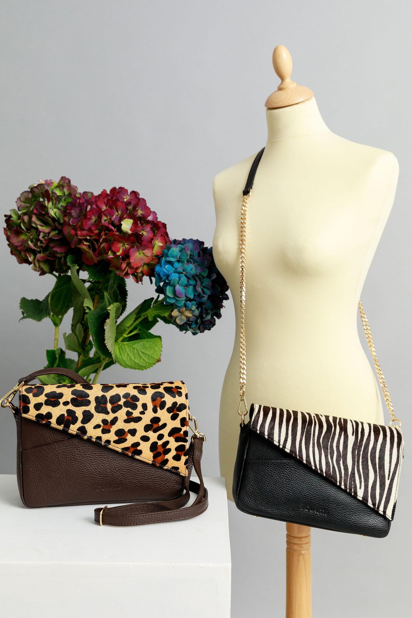 Chocolate brown and black crossbody bags with animal print flaps on a white surface with leather and chain straps attached. A bunch of three hydrangeas in burgundy, blue and purple are in the background.