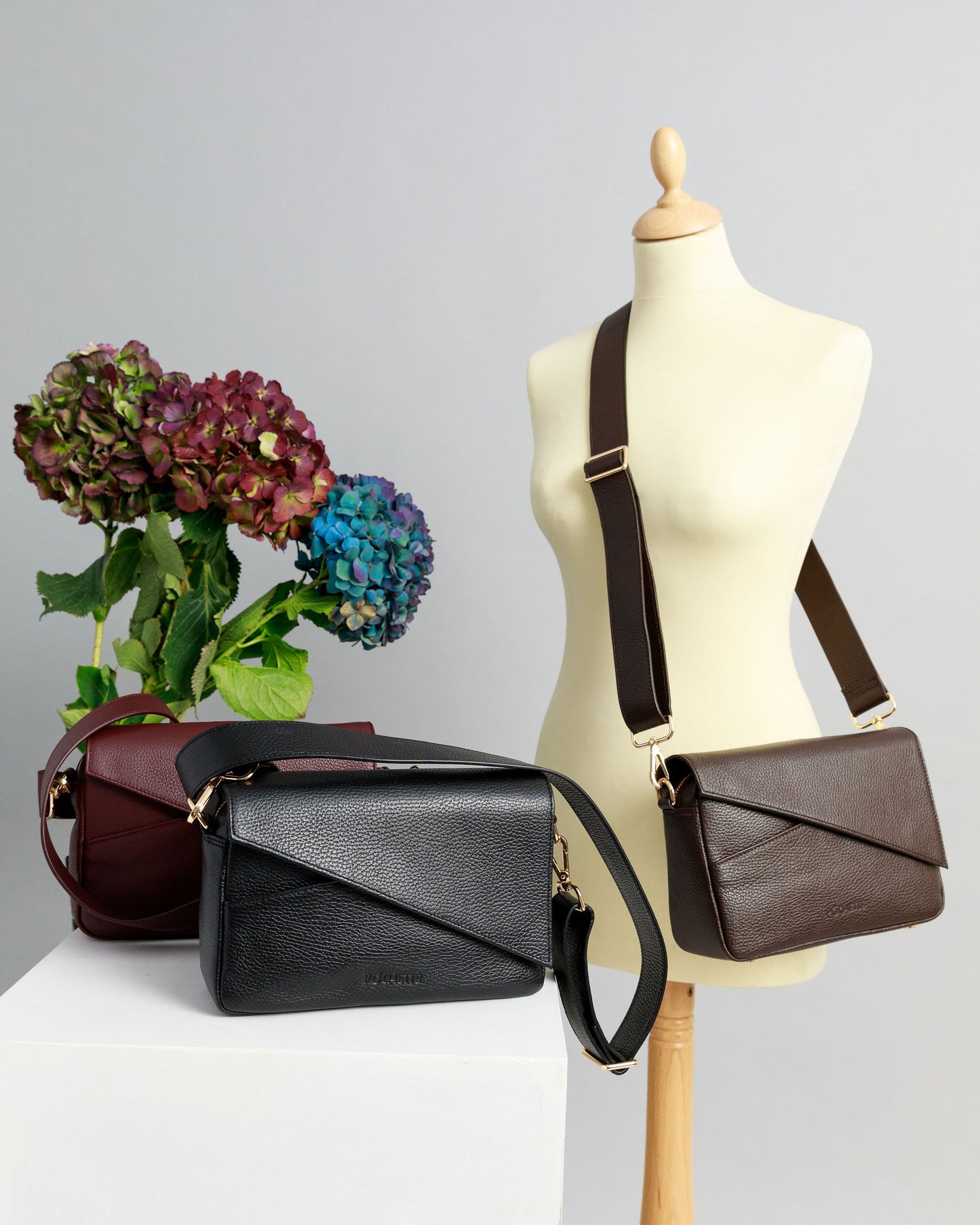 Burgundy and black crossbody bags on a white surface with wide leather straps attached. A chocolate brown crossbody bag is draped across a sewing mannequin, also with a wide leather strap. A bunch of three hydrangeas in burgundy, blue and purple are in the background.