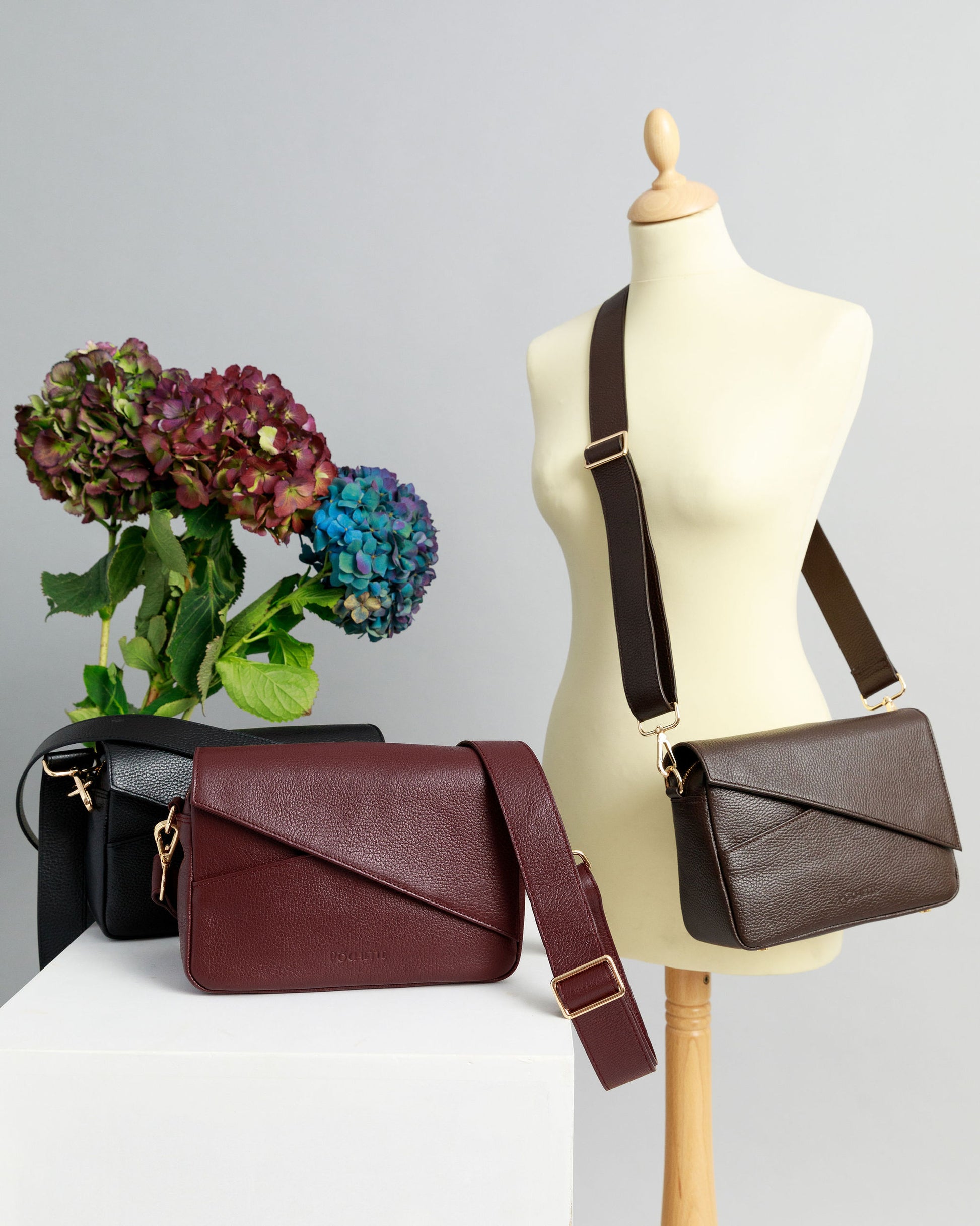 Chocolate brown and burgundy crossbody bags on a white surface with wide leather straps attached. A chocolate brown crossbody bag is draped across a sewing mannequin, also with a wide leather strap. A bunch of three hydrangeas in burgundy, blue and purple are in the background.