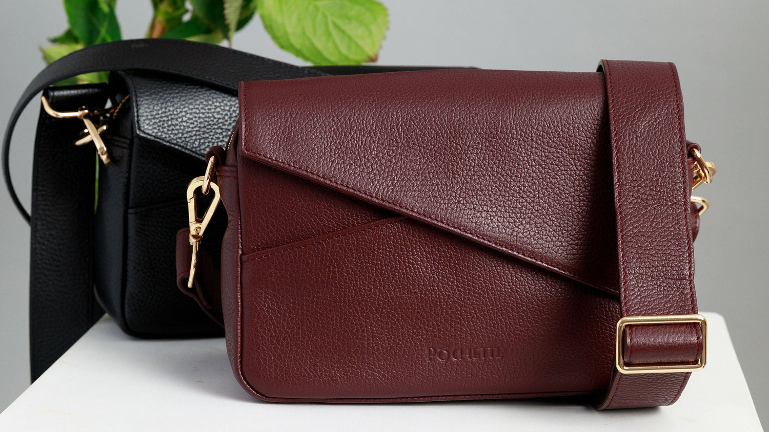 Chocolate brown and burgundy crossbody bags on a white surface with wide leather straps attached. A chocolate brown crossbody bag is draped across a sewing mannequin, also with a wide leather strap. A bunch of three hydrangeas in burgundy, blue and purple are in the background.