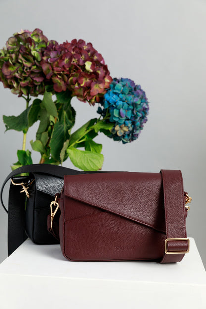 Chocolate brown and burgundy crossbody bags on a white surface with wide leather straps attached. A chocolate brown crossbody bag is draped across a sewing mannequin, also with a wide leather strap. A bunch of three hydrangeas in burgundy, blue and purple are in the background.