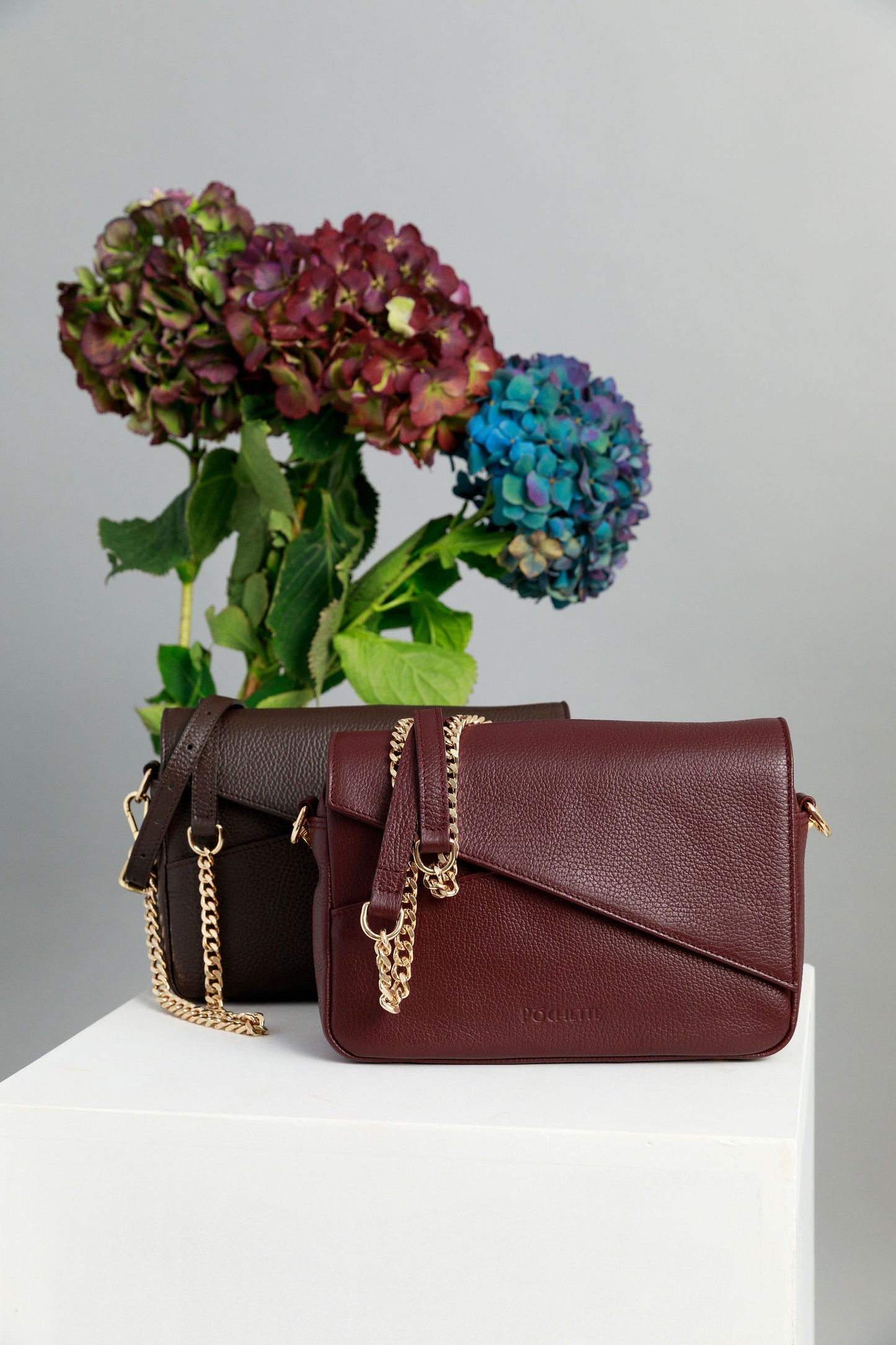 Chocolate brown and burgundy crossbody bags on a white surface with leather and chain straps attached. A bunch of three hydrangeas in burgundy, blue and purple are in the background.