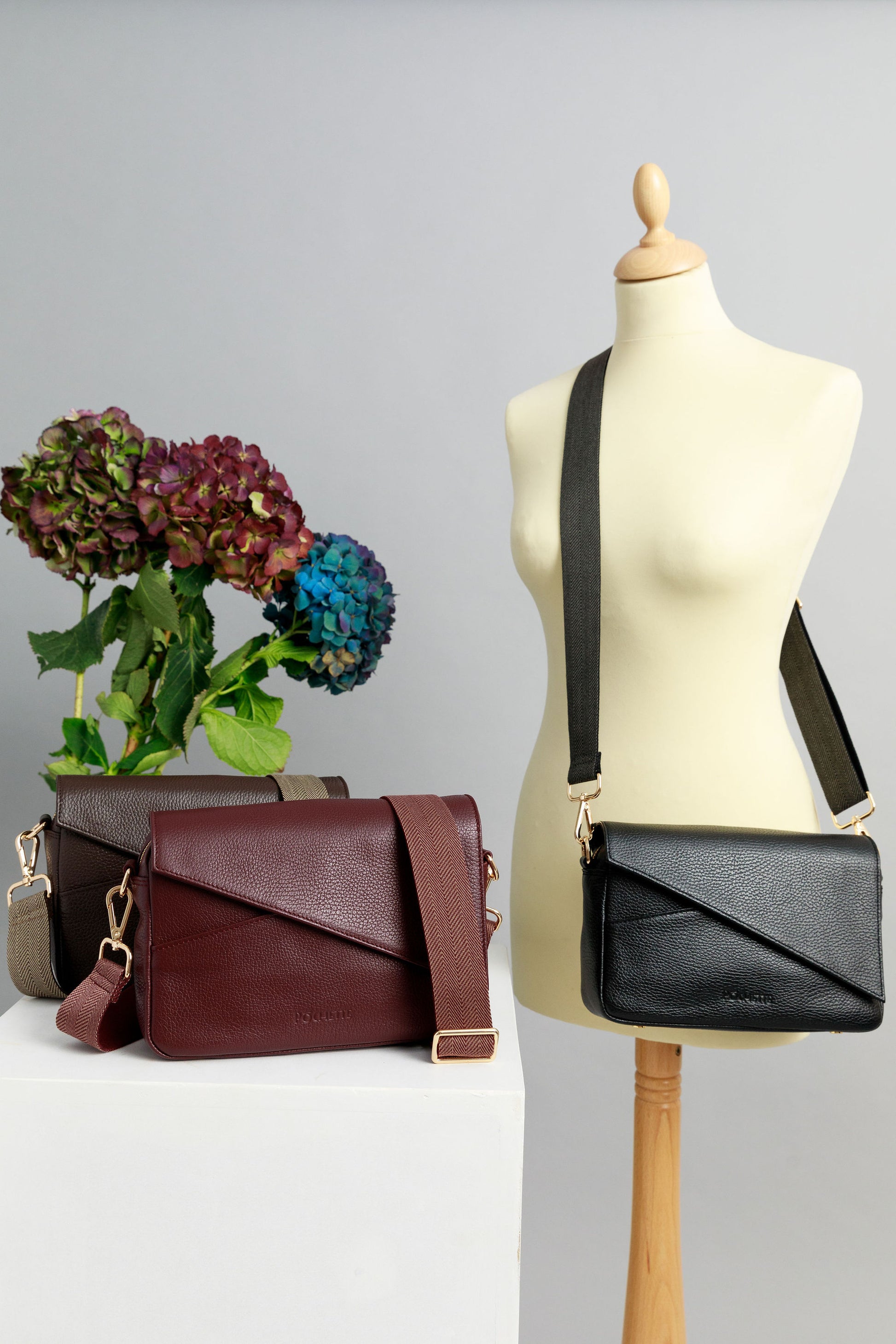 Chocolate brown and burgundy crossbody bags on a white surface with woven straps attached. A black crossbody bag is draped across a sewing mannequin with a striped woven strap. A bunch of three hydrangeas in burgundy, blue and purple are in the background.