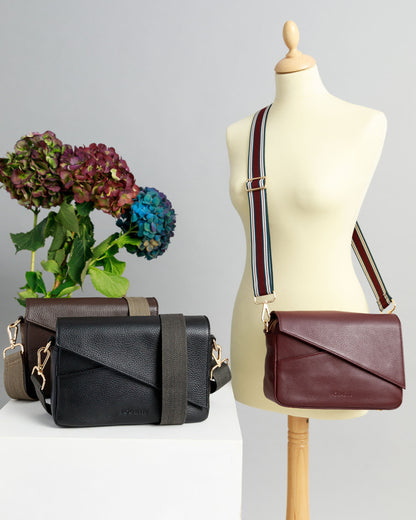 Chocolate brown and black crossbody bags on a white surface with woven straps attached. A burgundy crossbody bag is draped across a sewing mannequin with a striped woven strap. A bunch of three hydrangeas in burgundy, blue and purple are in the background.