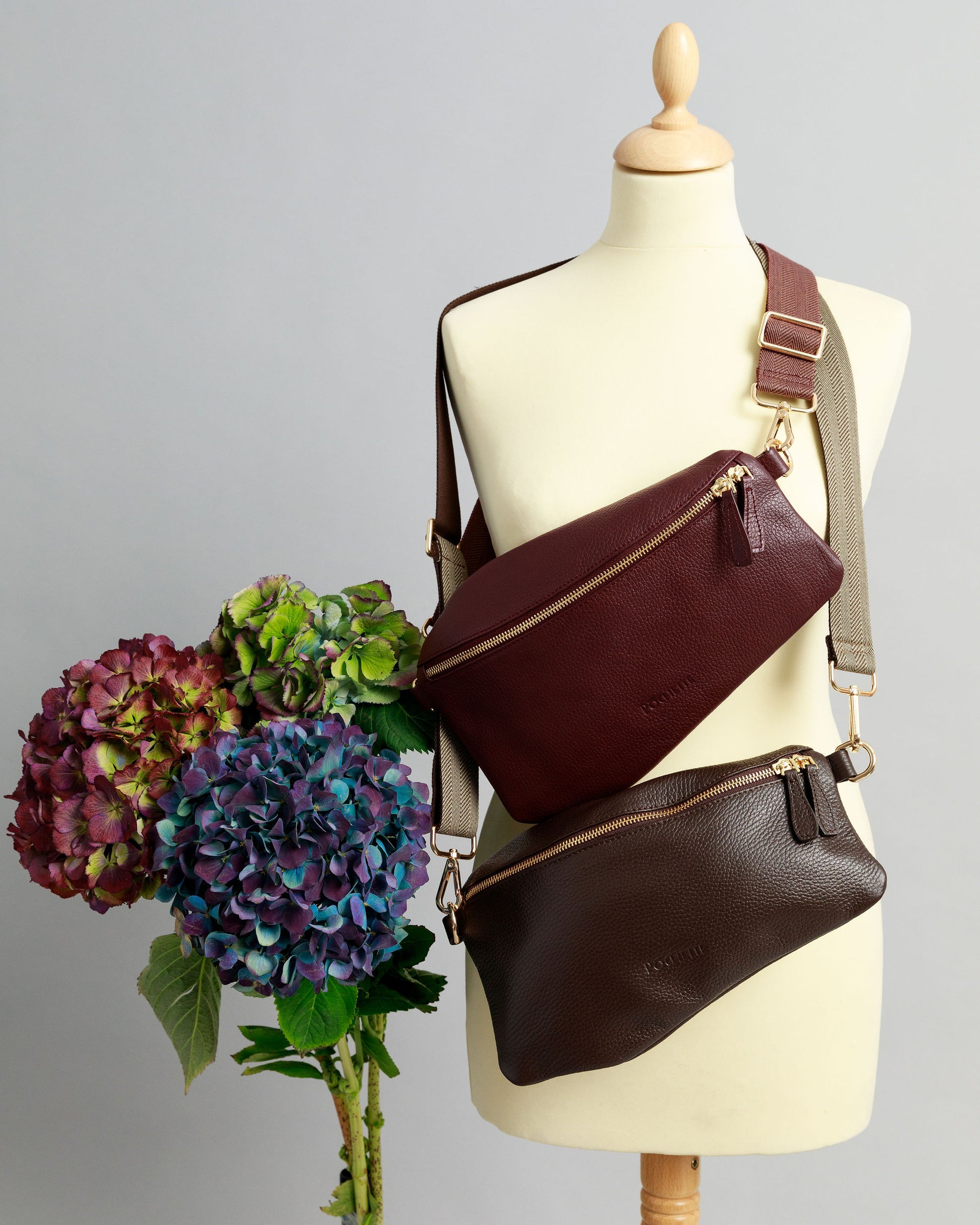 Burgundy and a chocolate brown Póca swing bags with woven crossbody straps on a sewing mannequin. A bunch of three hydrangeas in burgundy, blue and purple are beside it.