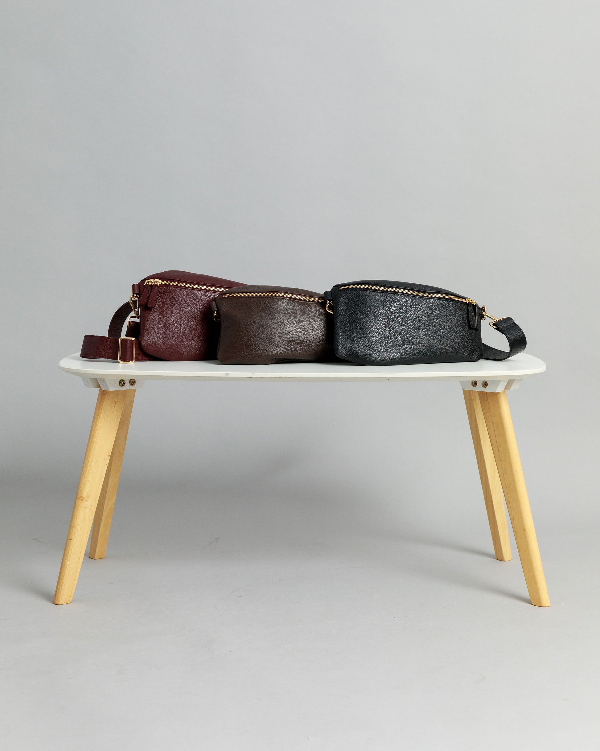 Burgundy, chocolate brown and black leather Póca swing bags on a white table with light coloured wooden legs on a grey background.