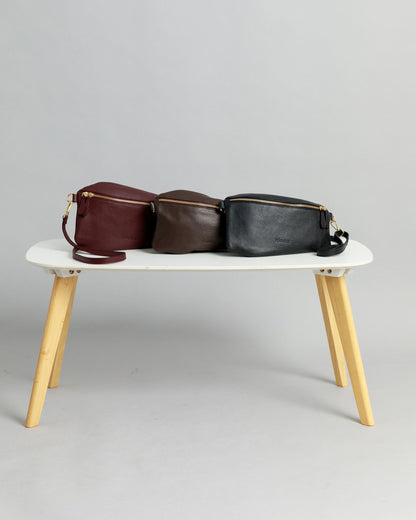 Burgundy, chocolate brown and black leather Póca swing bags on a white table with light coloured wooden legs on a grey background.