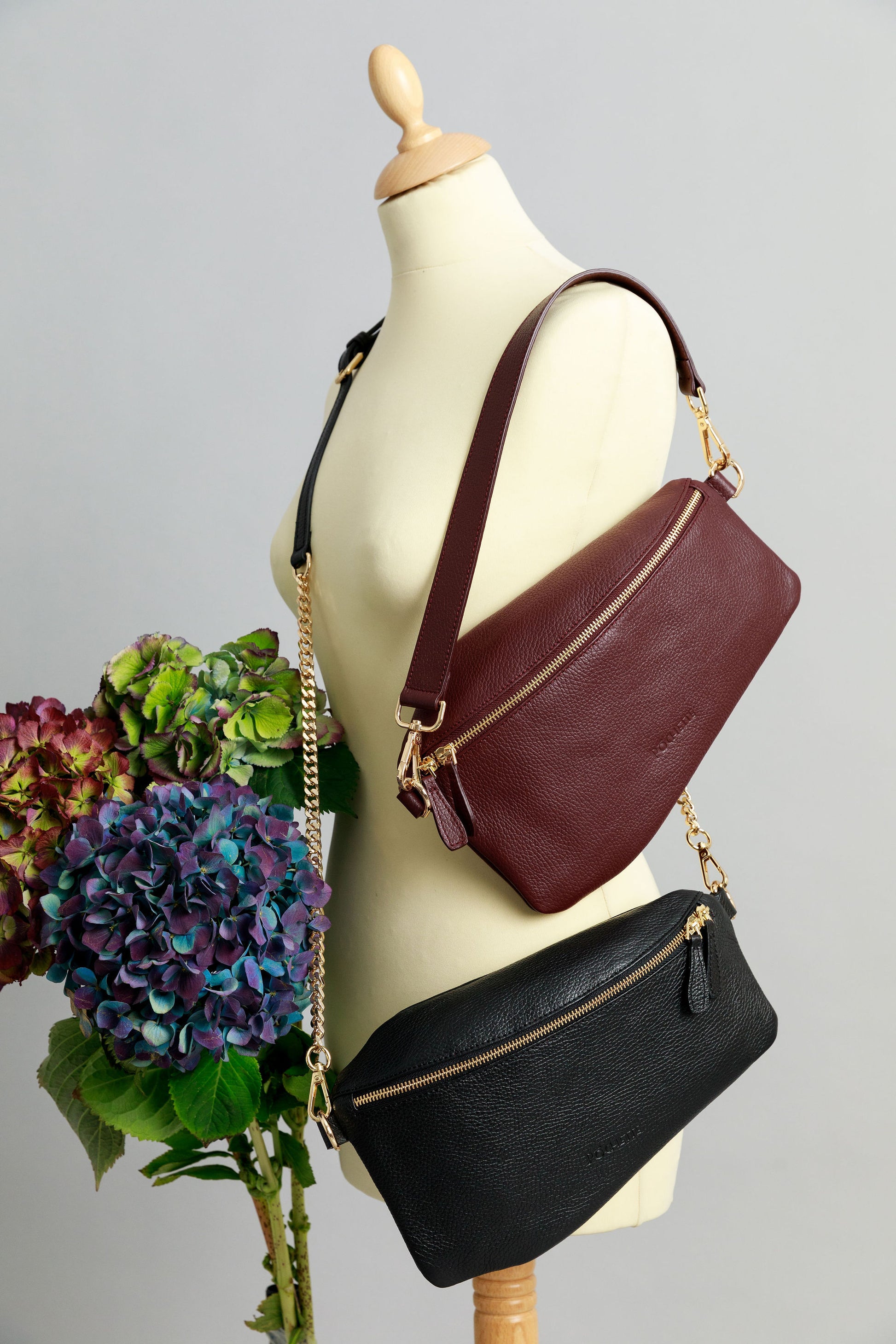 A burgundy Póca swing bag with a short leather shoulder strap and a black Póca swing bag with a leather and chain strap on a sewing mannequin. A bunch of three hydrangeas in burgundy, blue and purple are beside it.