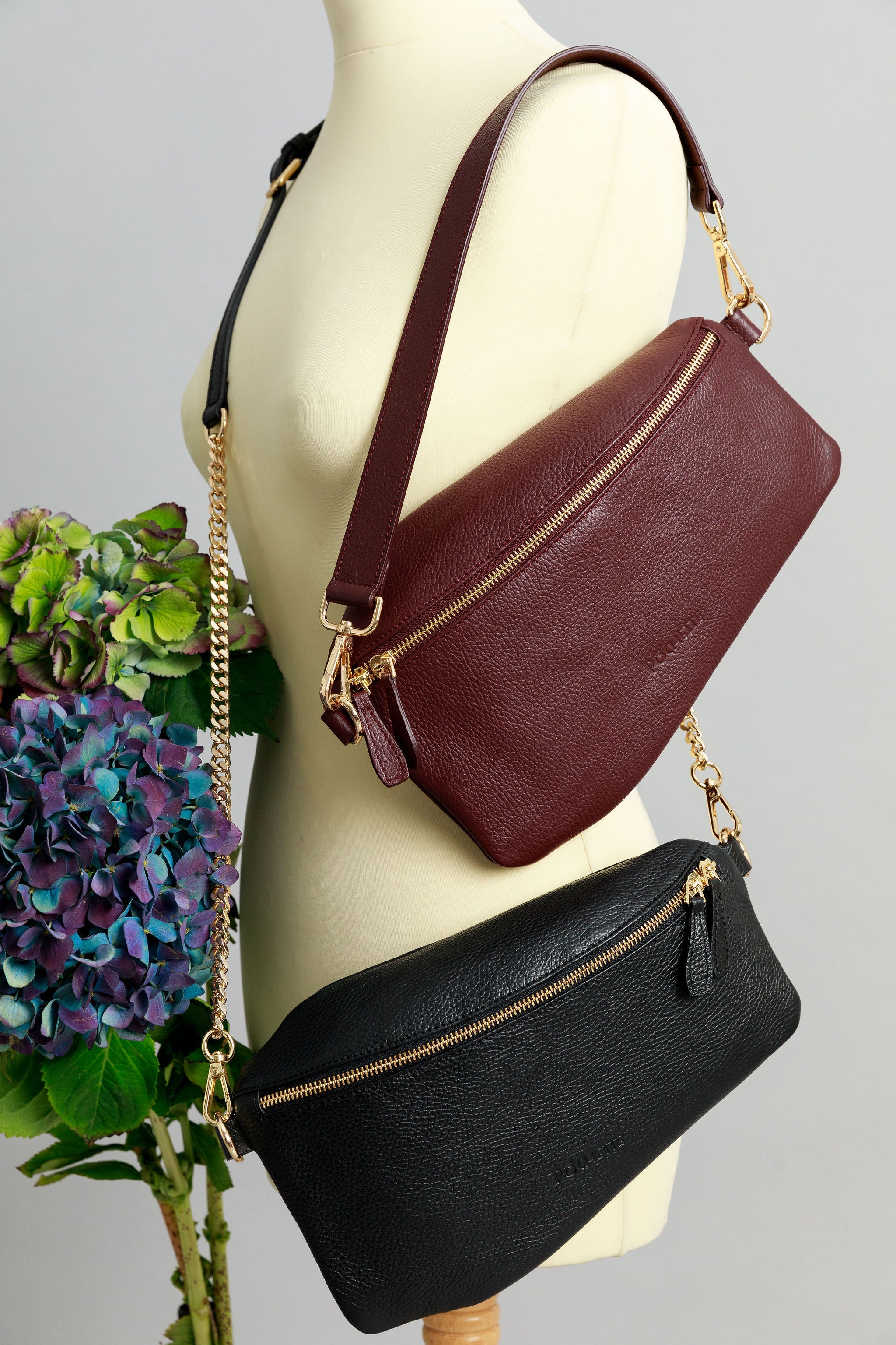 A burgundy Póca swing bag with a short leather shoulder strap and a black Póca swing bag with a leather and chain strap on a sewing mannequin. A bunch of three hydrangeas in burgundy, blue and purple are beside it.