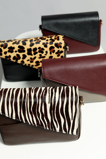 Four crossbody bags on a white surface - a burgundy crossbody bag with a black leather flap, a black bag with leopard print, a grey bag with a burgundy flap and a brown bag with a zebra print flap.