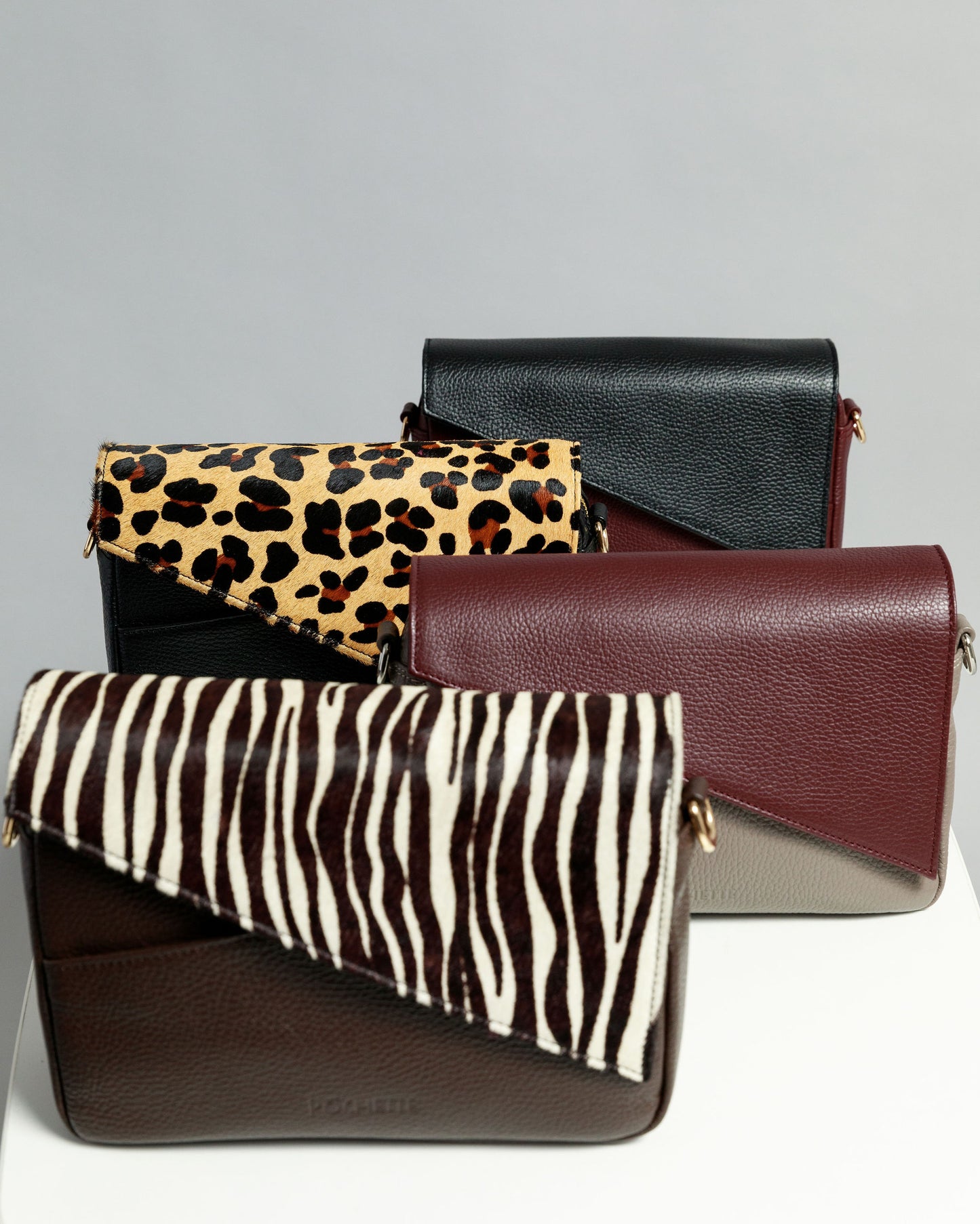 Four crossbody bags on a white surface with a grey background. From the back you can see a black leather flap on a burgundy crossbody bag, the next closest bag is black with a leopard print flap. This is followed by a pottery clay bag with a burgundy flap. At the front is a chocolate brown crossbody bag with a zebra print flap.