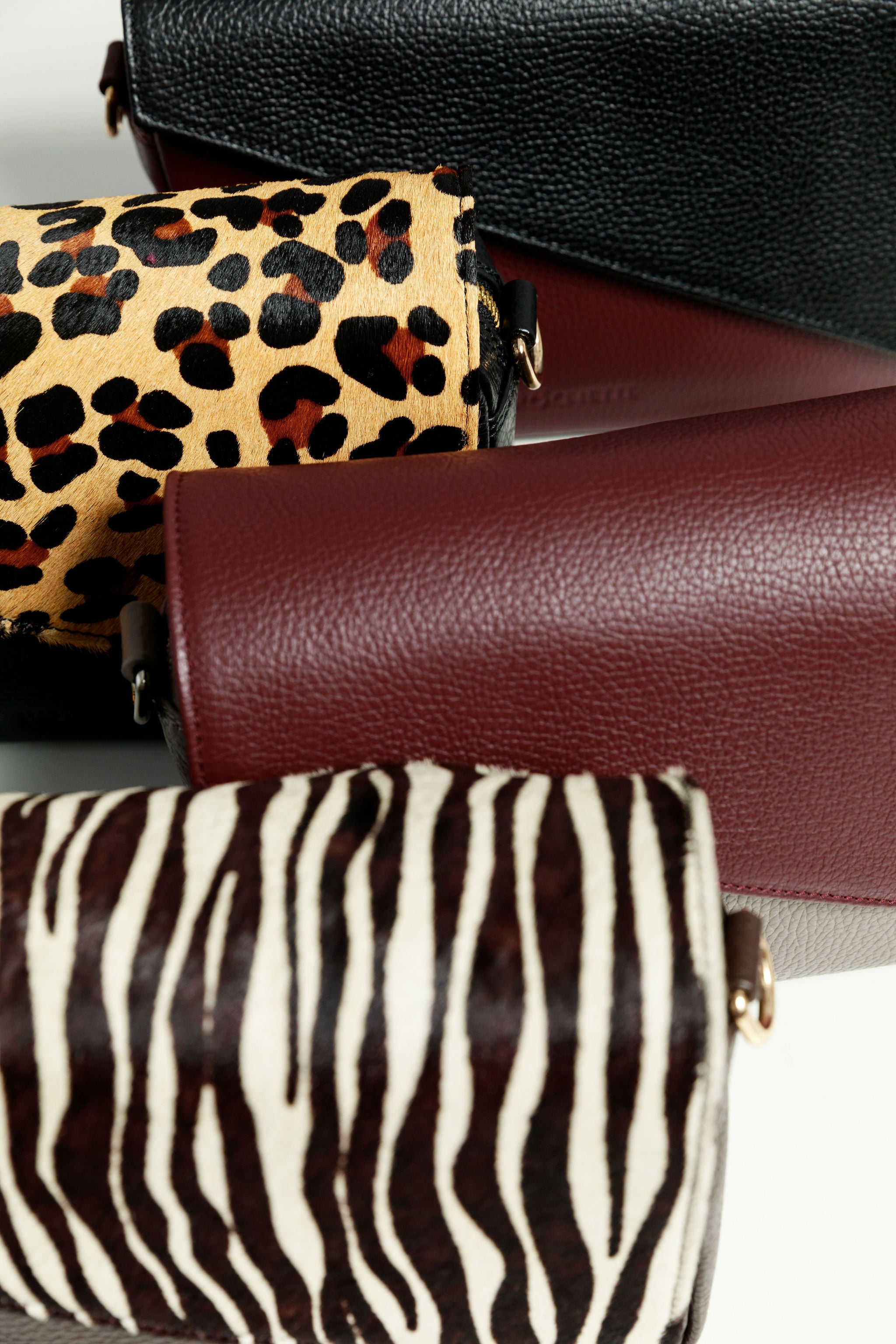 Four crossbody bags on a white surface - a burgundy crossbody bag with a black leather flap, a black bag with leopard print, a grey bag with a burgundy flap and a brown bag with a zebra print flap.