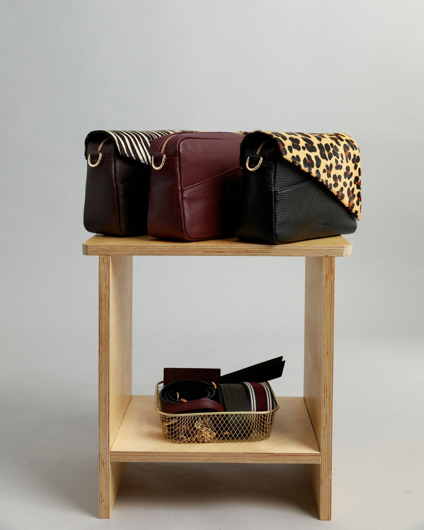 Three crossbody bags on a small plywood table with a small basket of accessories underneath infront of a grey background. On the left is a brown leather crossbody with a zebra print leather flap, followed by a burgundy crossbody bag and then a black crossbody bag with a leopard print flap.