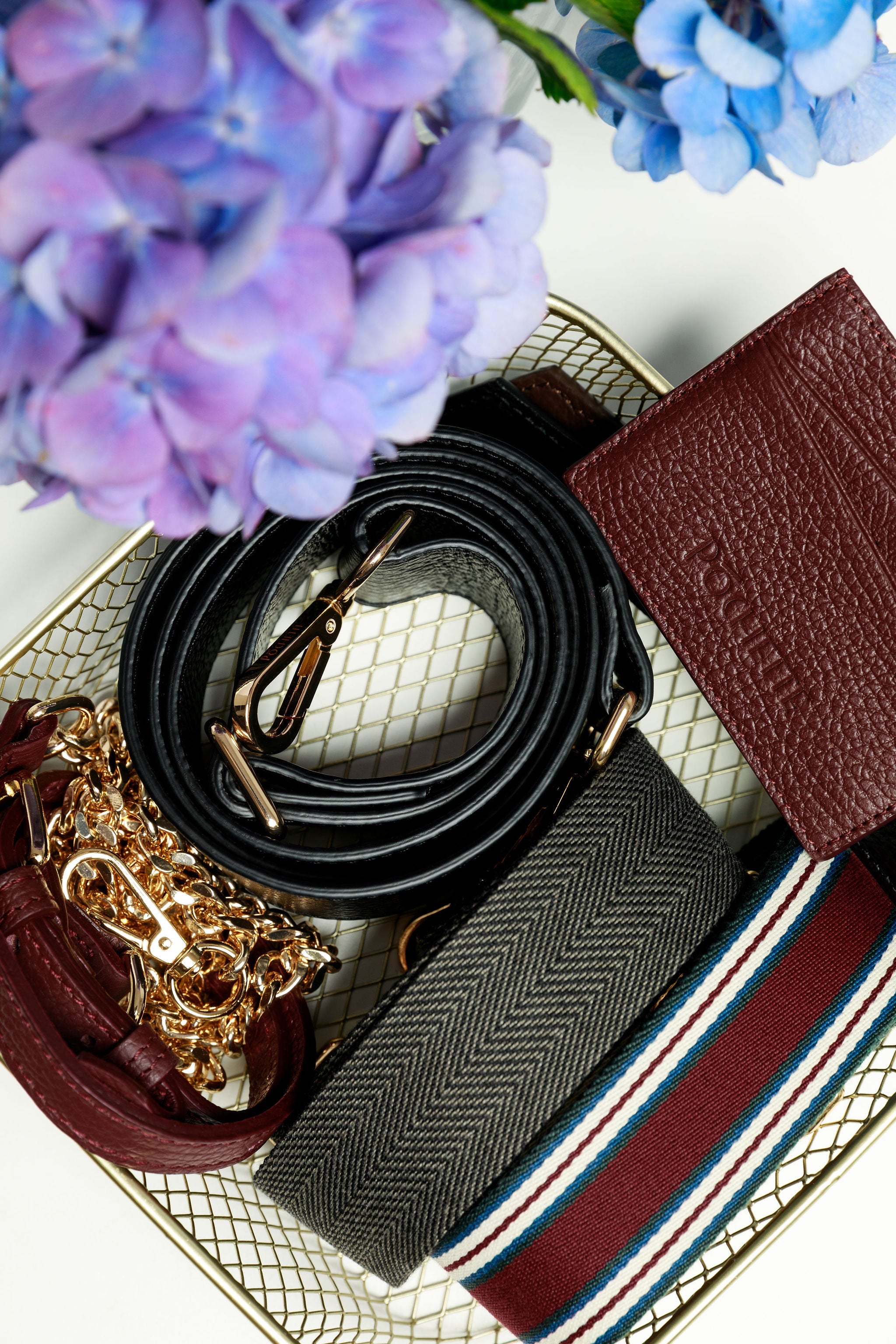 A collection of Póchette accessories in a wire basket including a burgundy wallet, a black woven strap, a striped woven strap, a burgundy chain and leather strap and a black wide leather strap. Blue and lilac hyrdrangeas are visible overhead.