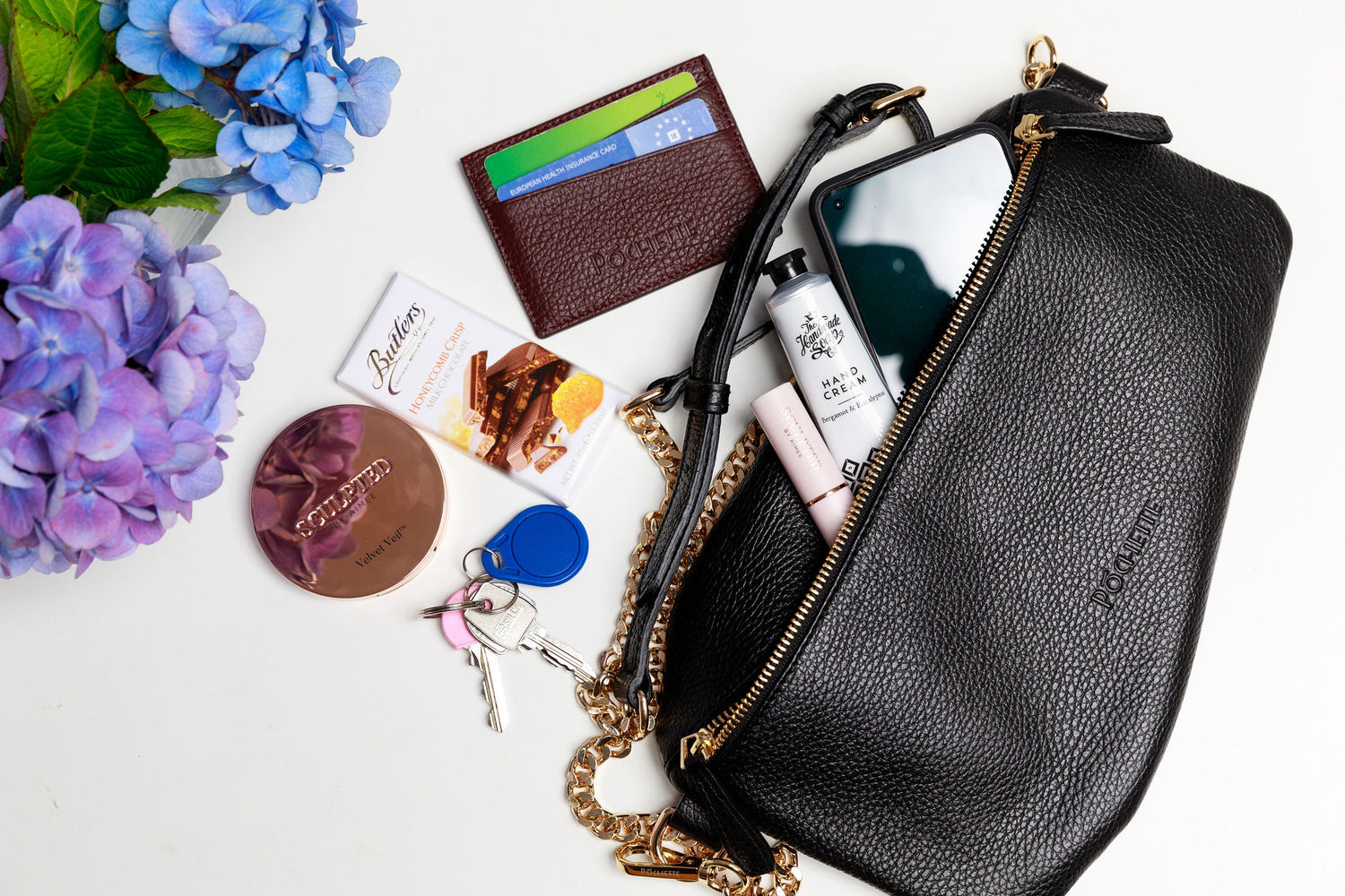 A black swing crossbody bag, called the Póca, on a white surface with blurred hydrangeas in the foreground. The contents of the bag are shown to be spilling out of the handbag, which can fit a mobile phone, handcream from the Homemade Soap Company, lipstick and face powder from Sculpted by Aimee, a burgundy Póchette wallet and some Butler's chocolate for emergencies. 