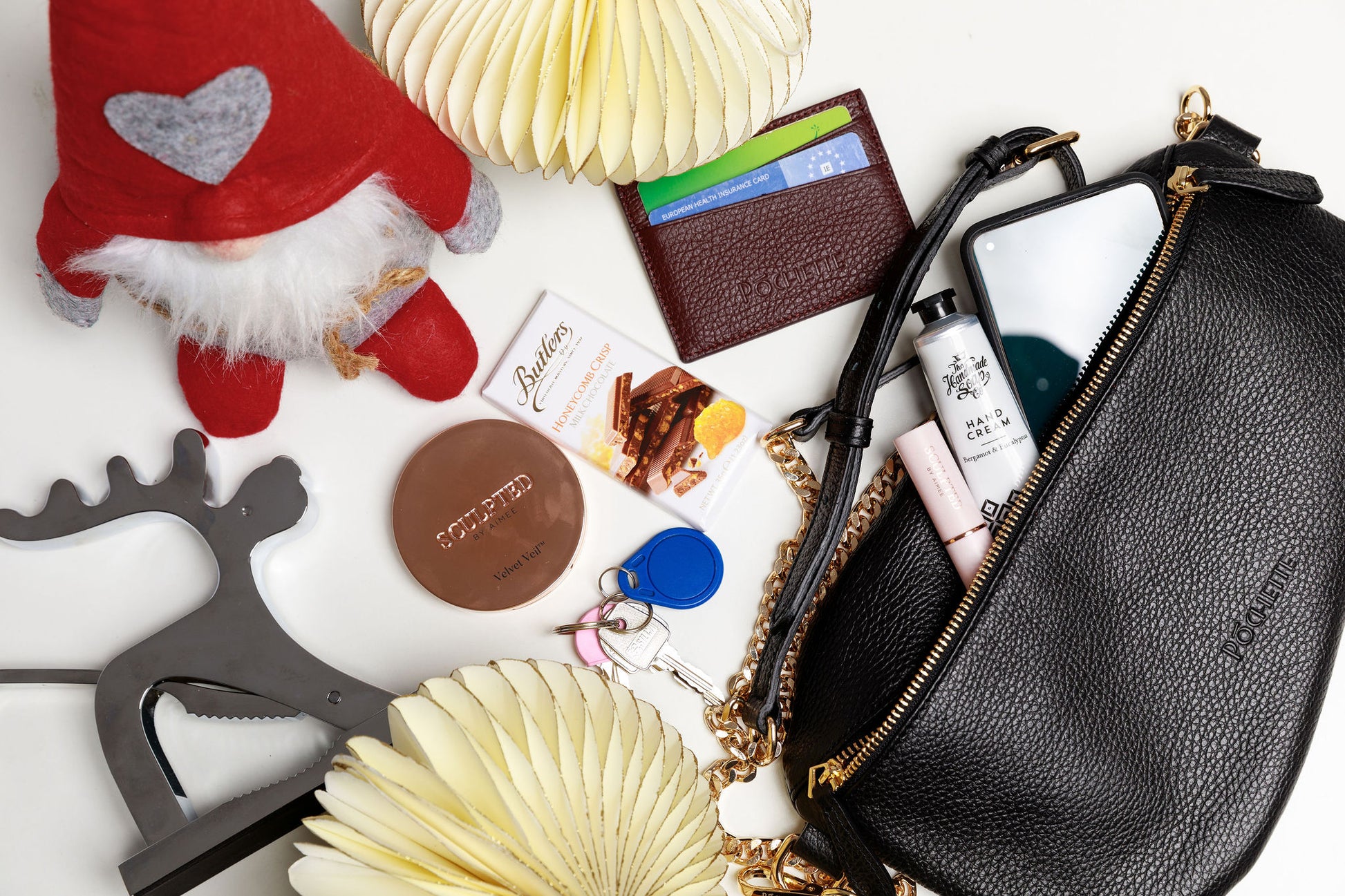 A black swing crossbody bag, called the Póca, on a white surface with christmas decorations. The contents of the bag are shown to be spilling out of the handbag, which can fit a mobile phone, handcream from the Homemade Soap Company, lipstick and face powder from Sculpted by Aimee, a burgundy Póchette wallet and some Butler's chocolate for emergencies. 