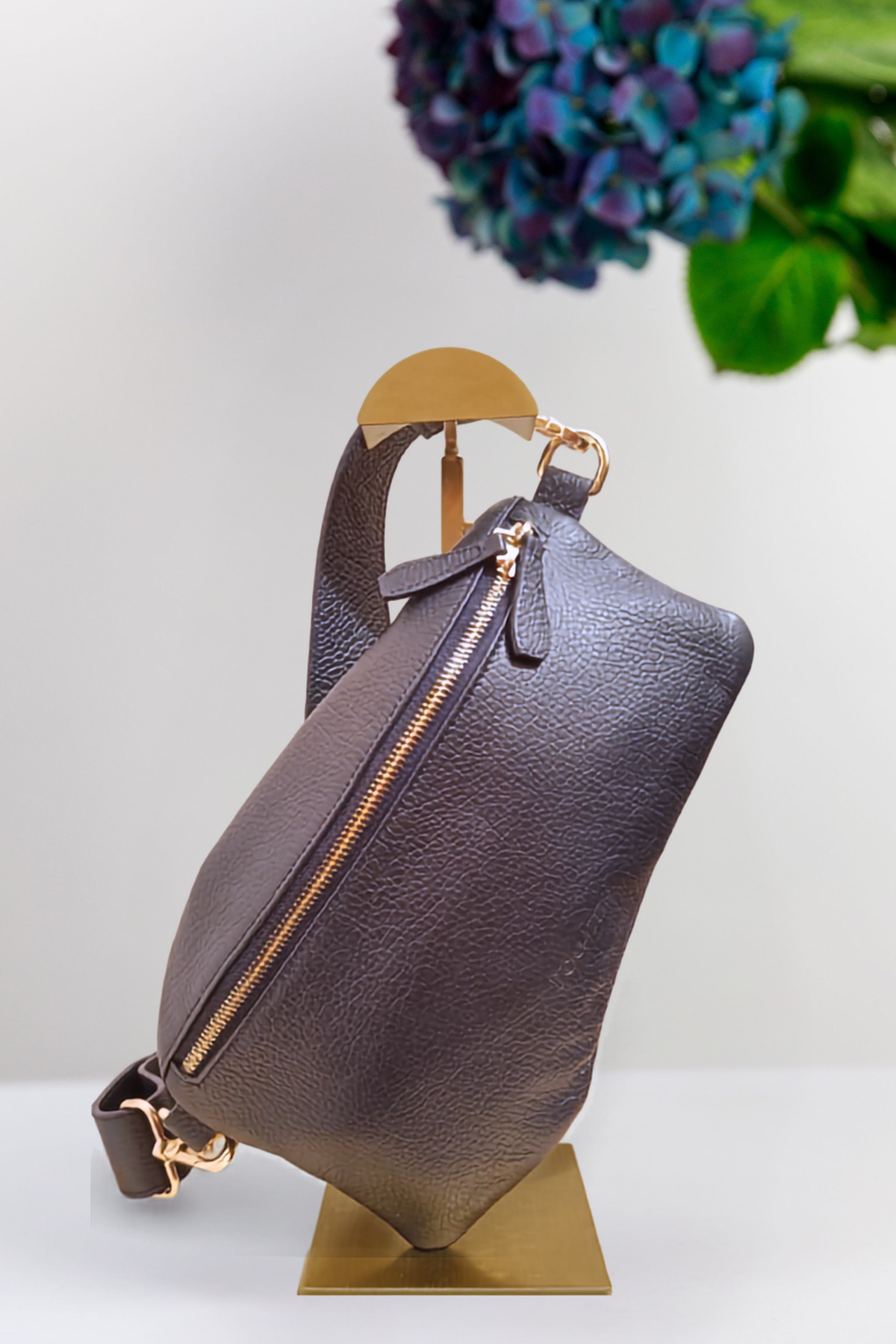 A navy pebbled leather handbag with gold-tone hardware and a wide leather strap, displayed on a gold-colored metal stand against a white background with a blurred purple flower in the upper right corner.