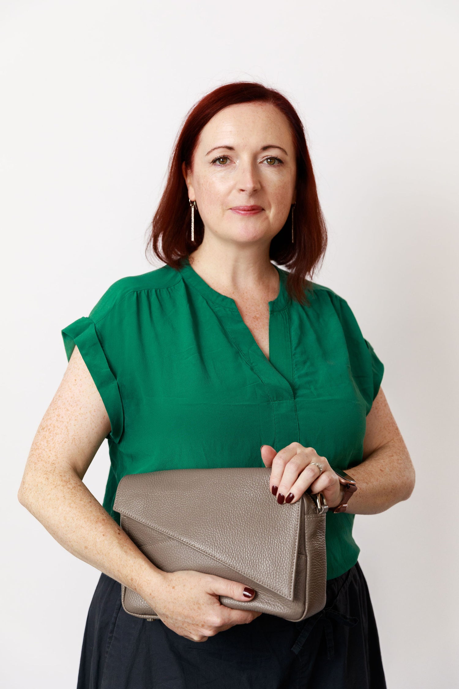 A photo of a woman with dark red hair and pale skin with some freckles wearing a green silk blouse, navy wide pants and silver drop-down earrings. She is proudly holding a pottery grey crossbody bag in her hands.