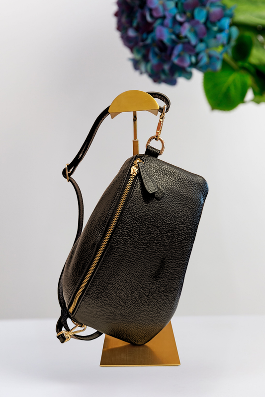 A black pebbled leather handbag with gold-tone hardware and a long narrow shoulder strap, displayed on a gold stand against a white background with blurred blue flowers in the upper right.