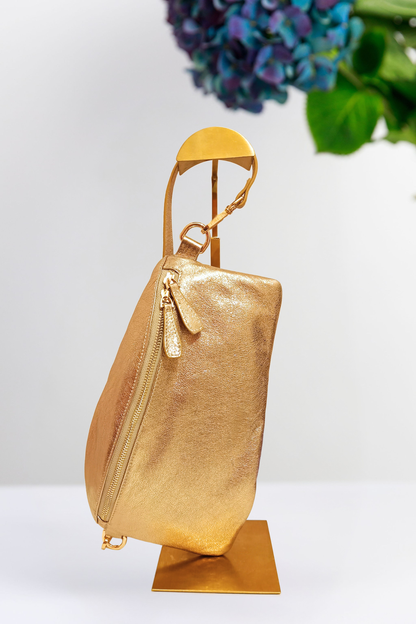 A metallic gold handbag displayed on a matching gold stand against a clean white background with a blurred floral arrangement in the upper right hand corner.