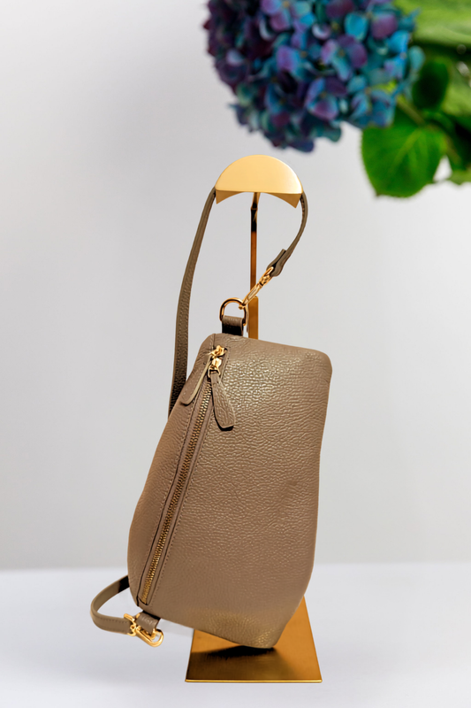 A grey pebbled leather shoulder bag with gold-tone hardware and a narrow leather strap, displayed on a gold-colored stand against a light gray background with a blurred purple flower in the upper right.