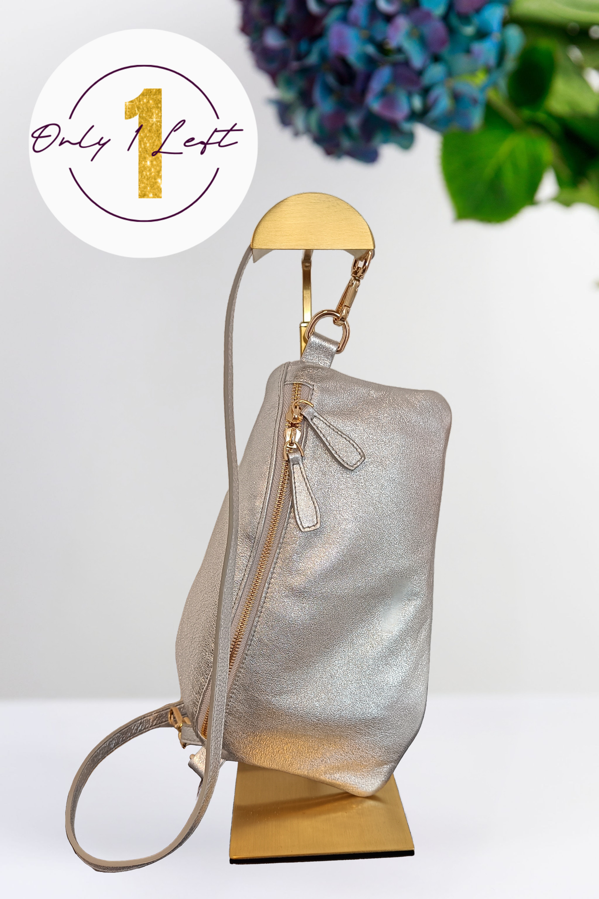 A metallic silver bag with gold-tone hardware, displayed on a gold stand against a white background with a floral prop in the upper right. A circular badge in the top left reads 'Only 1 Left' with a gold '1'.