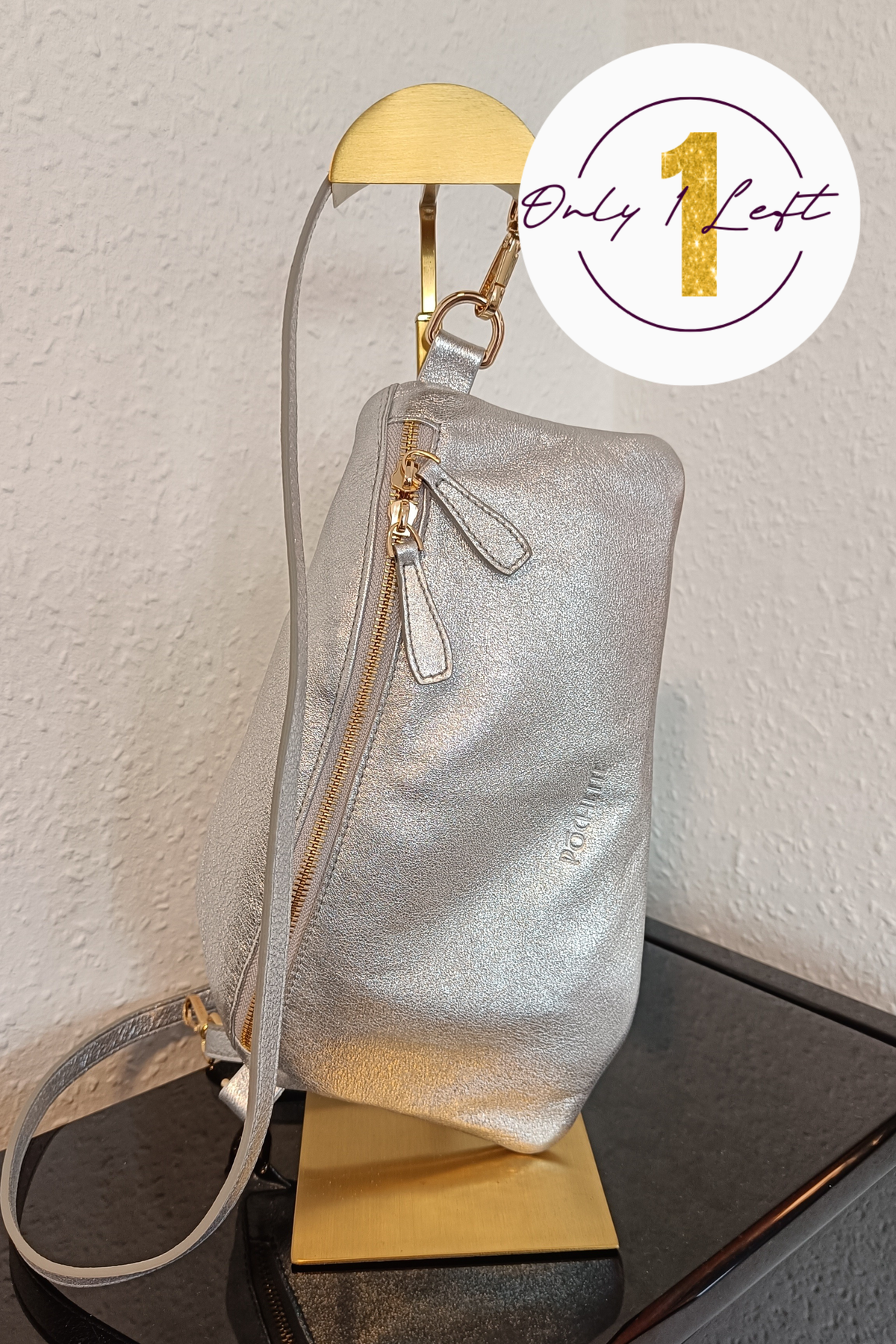 A metallic silver shoulder bag with gold-tone hardware, displayed on a gold stand against a textured off-white wall. A white circular badge in the top right corner features a gold '1' and the text 'Only Left'.