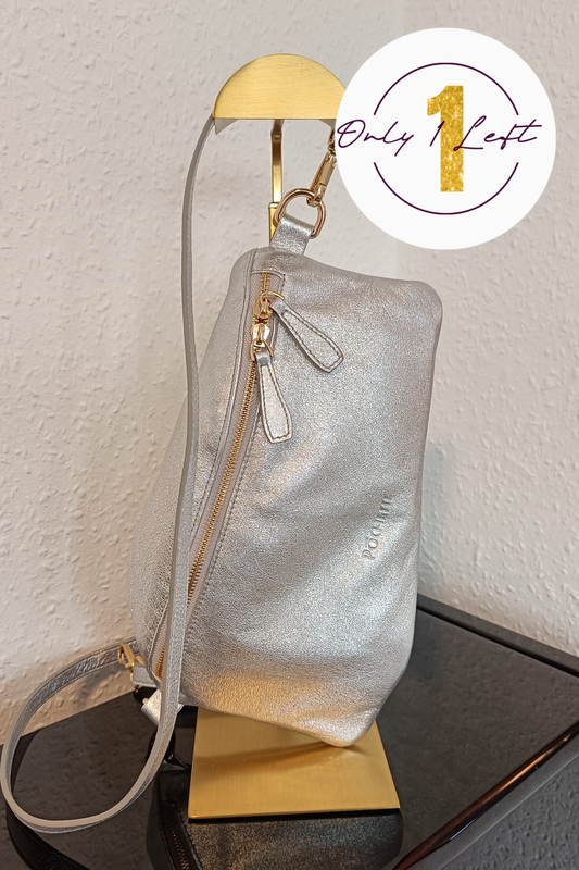 A metallic silver shoulder bag with gold-tone hardware, displayed on a gold stand against a textured off-white wall. A white circular badge in the top right corner features a gold '1' and the text 'Only Left'.