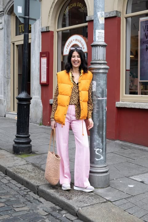 Photo of a dark haired white lady on the street wearing a leopard print clouse, yellow gilet, pink jeans and runners She's holding a straw tote bag.