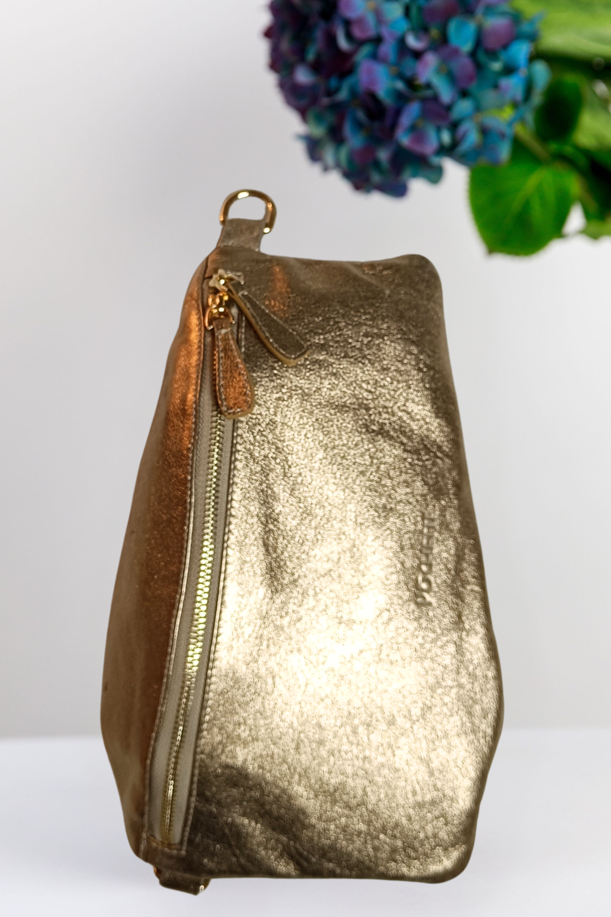 Gold metallic handbag with a zipper on a white background with a purple flower in the corner.