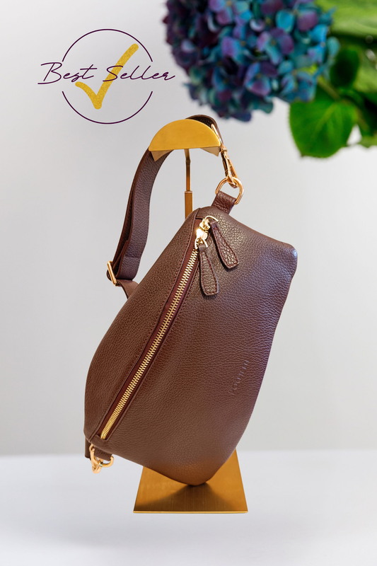 A burgundy pebbled leather shoulder bag with gold-tone hardware and a wide leather strap, displayed on a gold stand against a white background with a 'Best Seller' graphic overlay in the top left and flowers in the top right.
