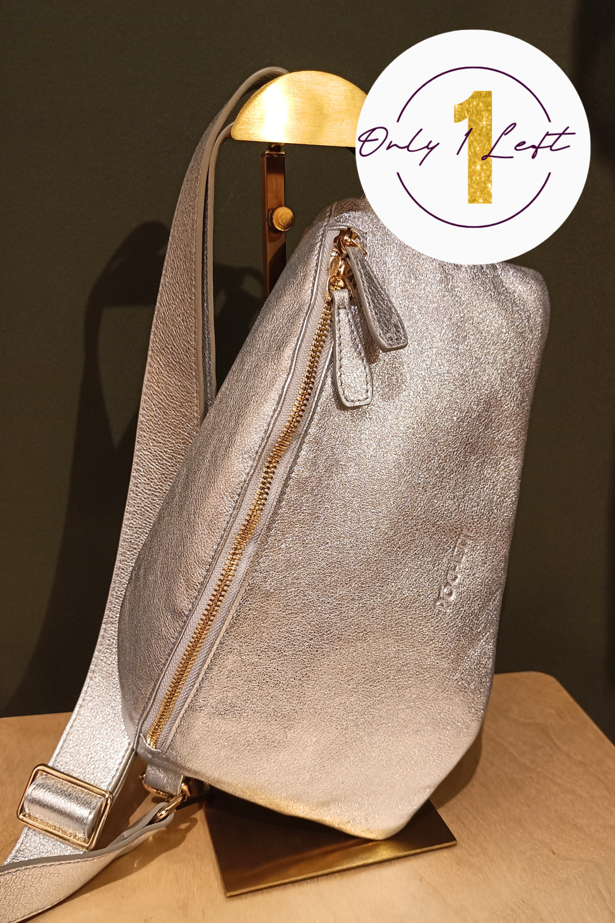 A metallic silver handag with gold-tone zippers, displayed on a gold stand against a dark green background. A white circular badge in the top right corner features a gold '1' and the text 'Only 1 Left'.
