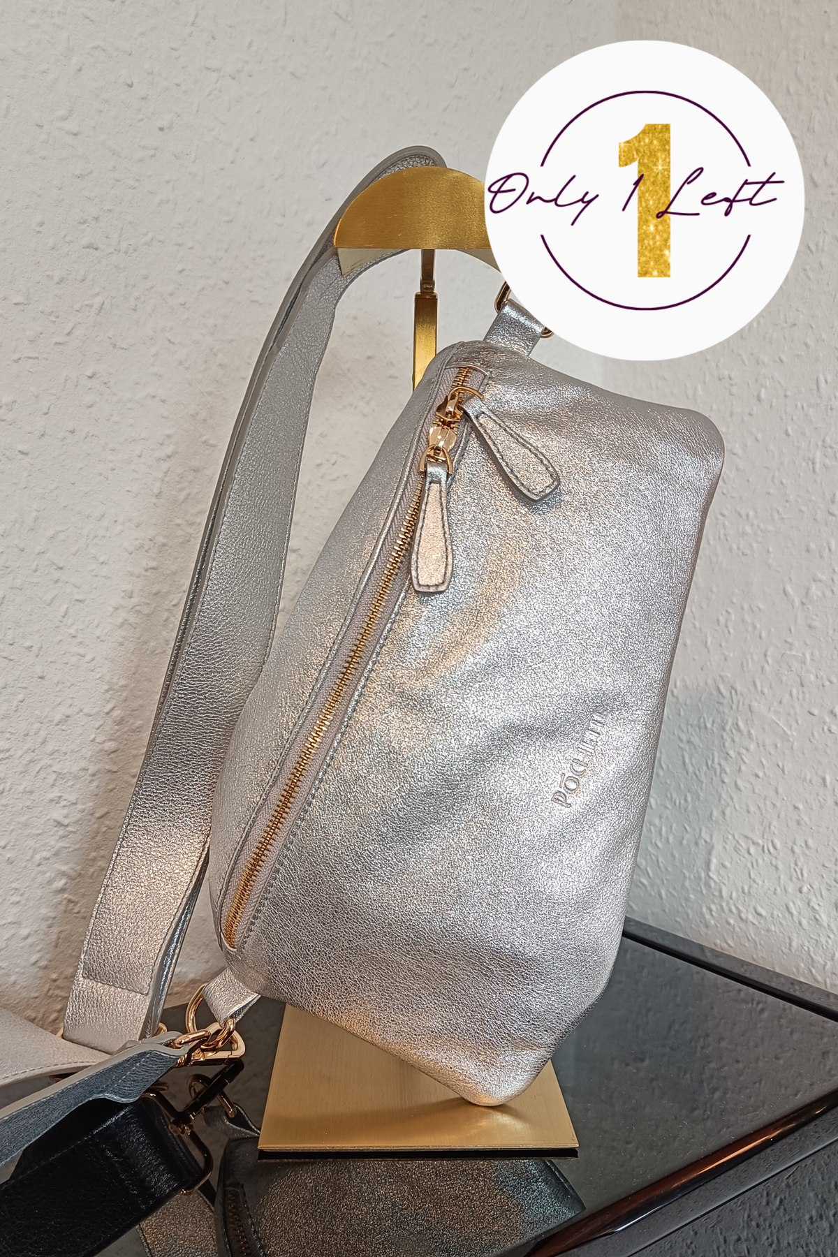 A metallic silver handag with gold-tone zippers, displayed on a gold stand against a white wall. A white circular badge in the top right corner features a gold '1' and the text 'Only 1 Left'.