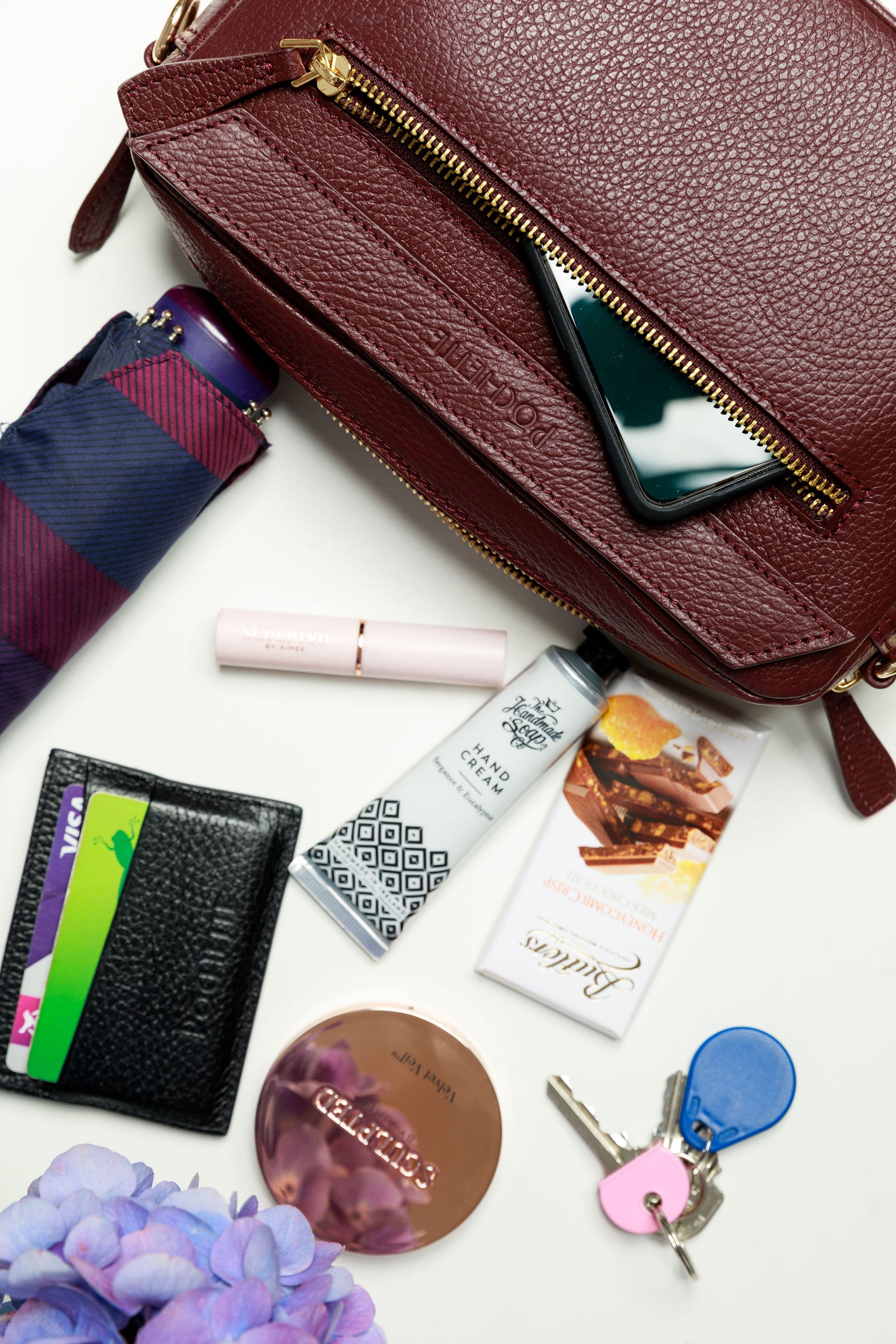 A burgundy crossbody bag on a white surface with blurred hydrangeas in the foreground. The contents of the bag are shown to be spilling out of the handbag, which can fit a mobile phone, handcream from the Homemade Soap Company, lipstick and face powder from Sculpted by Aimee, a black Póchette wallet, keys, an umbrella and some Butler's chocolate for emergencies. 